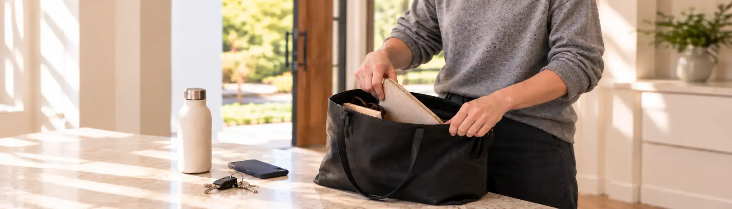 Person placing a small zip pouch into a tote bag on a kitchen bench beside keys, a phone and a water bottle.